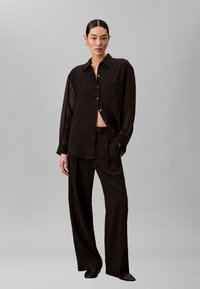 Sort, gennemsigtig skjorte med knapper, lange ærmer og en forlommer, kombineret med sorte bukser med vide ben og flade sorte sko.