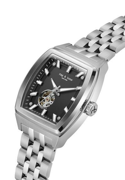 Hõbedane roostevabast terasest kell, millel on ruudukujuline must nägu, Rooma numbrid, nähtav tourbillon ja osaliselt lingiline metallist käevõru.