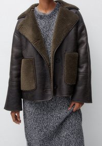 Chaqueta de piel marrón con cuello de piel sintética verde oliva y bolsillos frontales, con cierre de botón y mangas anchas. Suéter de punto gris texturizado debajo.