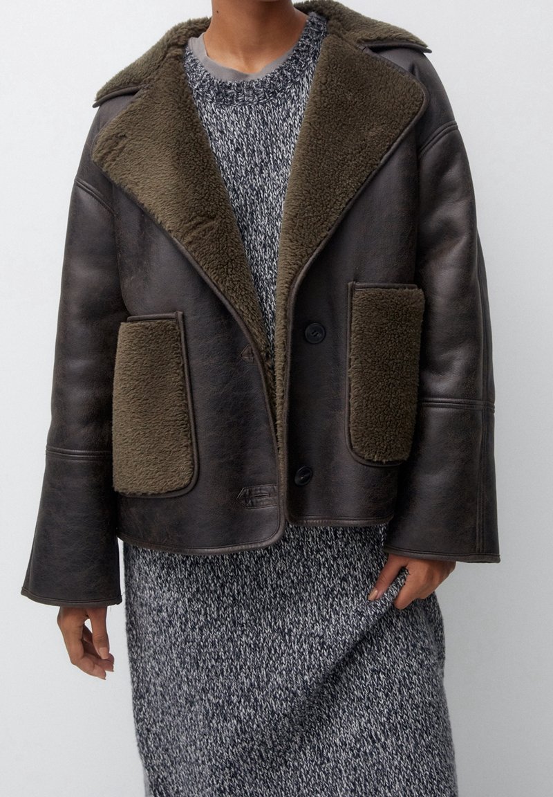 Chaqueta de piel marrón con cuello de piel sintética verde oliva y bolsillos frontales, con cierre de botón y mangas anchas. Suéter de punto gris texturizado debajo.
