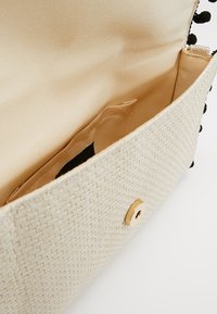 Woven beżowa torebka kopertówka z złotymi akcentami, z wewnętrzną kieszonką na suwak oraz gładkim kremowym wykończeniem. Krawędzie zdobione są czarnymi pomponami.