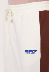 Balti ir rudi sportiniai kelnes su raiščiu juosmenyje, šonine kišene ir mėlynu EA7 Emporio Armani logotipo ženklu kairėje koja.