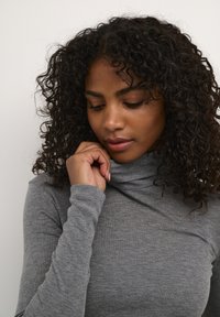 Grau geschnittener Rollkragenpullover mit langen Ärmeln, aus weichem, strukturiertem Stoff. Der Kragen ist umgeschlagen, und die Passform wirkt eng anliegend. Lockige Haare sind sichtbar.