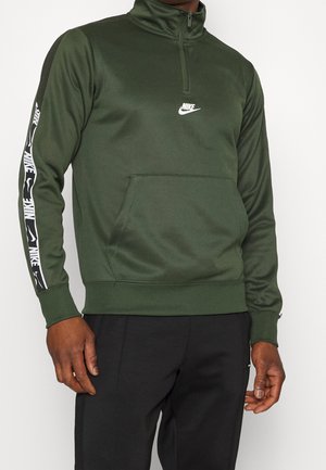 Homme portant un sweat-shirt Nike vert olive à fermeture éclair quart, avec un logo blanc sur la poitrine et des bandes de marque sur les manches, associé à un pantalon noir.