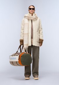 Casaco de fleece bege, cachecol castanho, mala de palha verde-oliva e laranja com logótipo, calças largas castanhas e sapatos plataforma com detalhes metálicos.