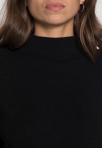 Svart stickad tröja med hög hals, med ribbad textur och långa ärmar. Materialet verkar mjukt och flexibelt.