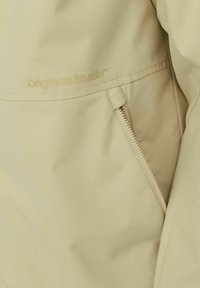 Lys beige jakke med en glat tekstur, en lynlåslomme og det præget logo "Originals Studio™" på stoffet.