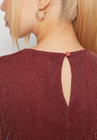 Bordeaux gekleurde top met textuur en verticale ribbels, voorzien van een sleutelgat uitsnijding aan de achterkant, bevestigd met een kleine rode knop. Zichtbaar zilveren ringoorbel.
