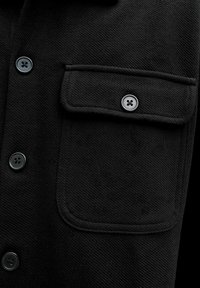 Czarna kurtka z zapięciem na guziki z przodu, dużą kieszenią na piersi z klapką oraz teksturowanym materiałem o subtelnym skośnym splocie.