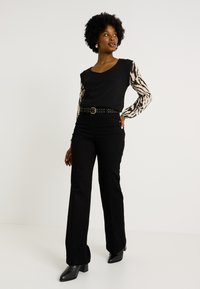 Top negro con mangas de estampado animal, combinado con jeans de campana negros de cintura alta y botas de tobillo negras con tacón. Cinturón con detalles en dorado.