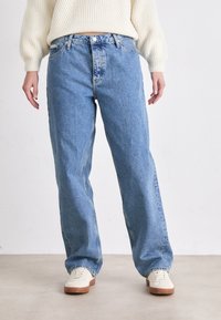 Világoskék farmernadrág egyenes szárú dizájnnal, öt zsebes stílussal és cipzáros záródással. Fehér sneaker cipőkkel párosítva, semleges háttér előtt.