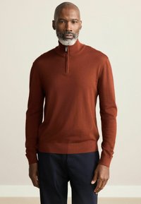 Rudai raudonos spalvos zip-up puloveris, pagamintas iš minkštos medžiagos, su ilgomis rankovėmis ir raštuotais rankogaliais. Yra pusės ilgio apykaklė ir prigludusi apačia.