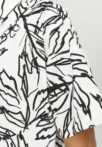 Chemise à manches courtes en tissu blanc avec un motif de feuilles abstraites noires ; elle présente un col et une coupe décontractée.