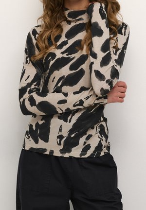 Femme portant un col roulé beige avec des taches abstraites noires et un pantalon noir, bras croisés, sur un fond uni.
