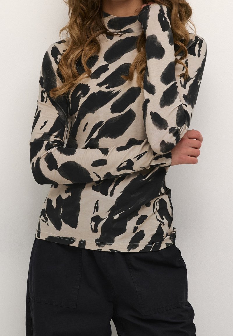 Femme portant un col roulé beige avec des taches abstraites noires et un pantalon noir, bras croisés, sur un fond uni.