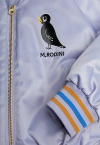 Lilac krūšu jaka ar melna putna grafiku un tekstu "M. RODINI". Iezīmētas ribatās aproces ar zilām un oranžām svītrām, zelta rāvējslēdzēja detaļa.