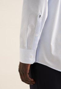 Camicia a righe blu chiaro e bianca con polsini abbottonati. Presenta un ricamo floreale sul polsino sinistro. Materiale in cotone con una texture liscia.