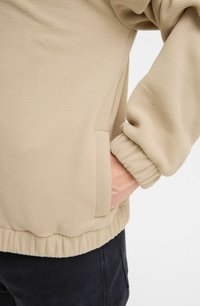 Beige fleece pullover med ribbede manchetter, elastisk kant og en sidelomme. Blød tekstur og afslappet pasform.