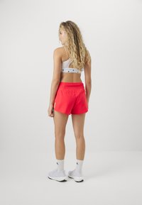 Pantalones cortos deportivos rojos con una textura suave, cinturilla elástica y un pequeño logo en el lateral. Combinados con un sujetador deportivo blanco y calcetines blancos.