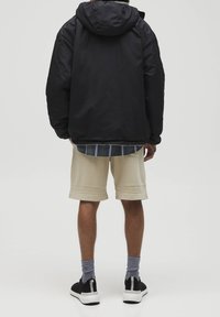 Czarna kurtka z kapturem, jasnobeżowe szorty, czarne buty, szare skarpetki, w dolnej części widoczna koszula w kratę, jednolite tło.