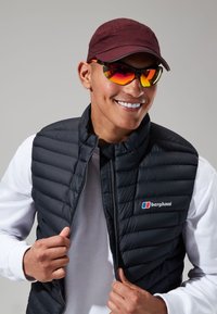 Zwarte puffer vest met een rits, met een logo. Daaronder een wit longsleeve shirt. Een bordeauxrode pet en spiegelende zonnebril maken de look compleet.