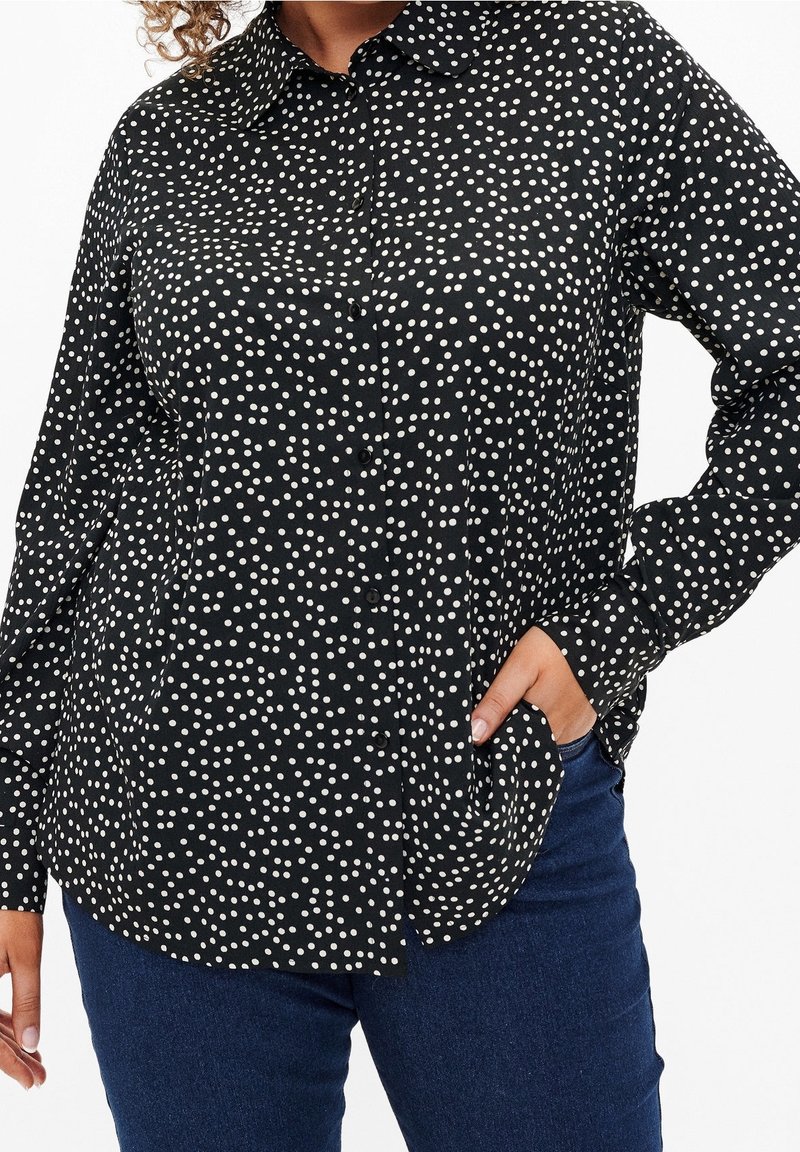 Chemise noire à manches longues avec des pois blancs portée avec un jean bleu, une main dans la poche et cheveux bouclés visibles.
