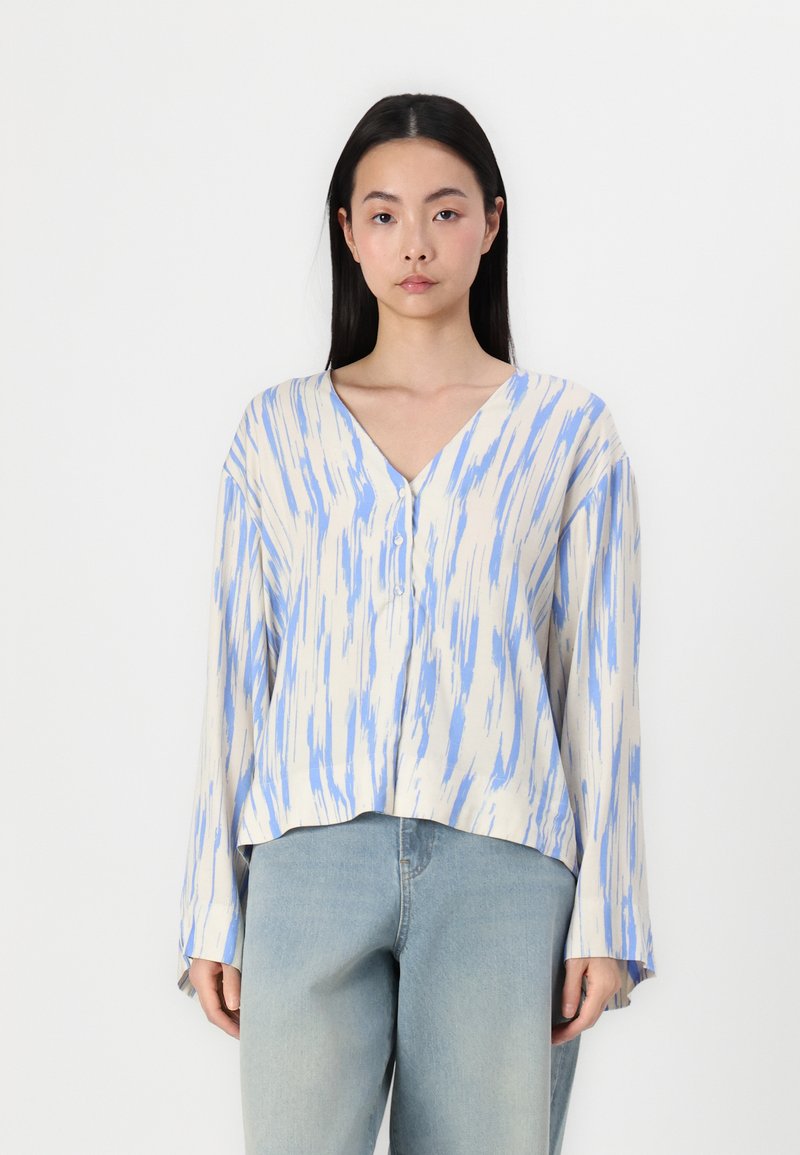 Leichte, V-Ausschnitt-Bluse mit blau-creamfarbigem abstraktem Muster, langen Ärmeln und Knopfdetails am Ausschnitt. Getragen mit hellblauen Jeans.