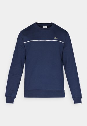 Sweatshirt bleu marine en tissu doux, doté d'une bande horizontale blanche, d'un patch logo sur la poitrine et de détails de marque sur les manches.