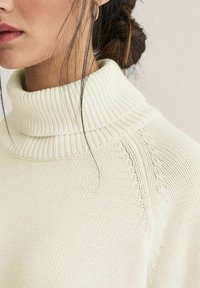 Neulepaita korkea kaulus kevyessä kermanvärissä, jossa on resori kaulus ja koristeellinen kuvio olkapäillä ja rinnassa. Pehmeä pinta.