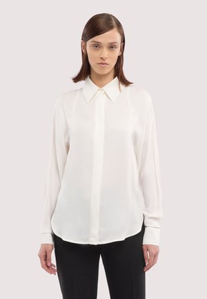 Kobieta o prostych brązowych włosach, ubrana w białą koszulę na guziki z długim rękawem oraz czarne spodnie, stojąca na tle jasnego, jednolitego tła.