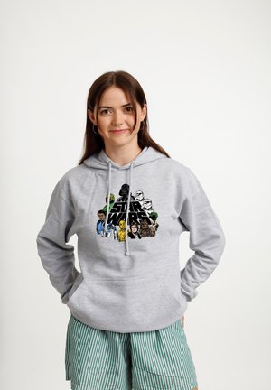 Młoda kobieta o brązowych włosach, nosząca kolczyki koła, szarą bluzę z kapturem Star Wars oraz zielone, prążkowane szorty, stoi z rękami za plecami.