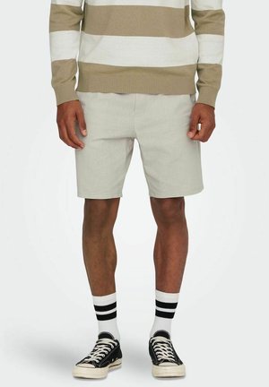 Mann trägt hellgraue Shorts, beige-weiß gestreiften Pullover, weiße Socken mit schwarzen Streifen und schwarze Sneaker mit niedrigem Schaft.