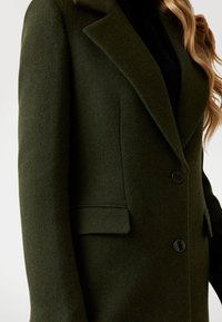 Blazer din lână verde închis, cu o suprafață texturată, rever ascuțite și două buzunare frontale, închise cu butoane negre.