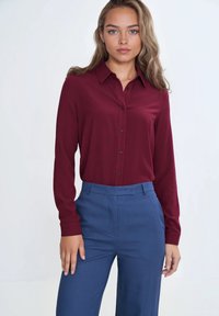Burgundowa koszula z długim rękawem i kołnierzykiem, zestawiona z niebieskimi, dopasowanymi spodniami. Materiał wydaje się gładki i lekko błyszczący.