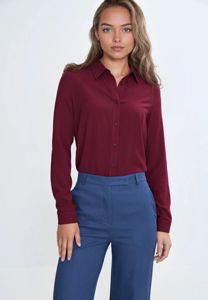 Burgundowa koszula z długim rękawem i kołnierzykiem, zestawiona z niebieskimi, dopasowanymi spodniami. Materiał wydaje się gładki i lekko błyszczący.
