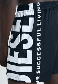 Black swim shorts featuring large white text along the side. Made from lightweight material with an elastic waistband and a smooth texture.