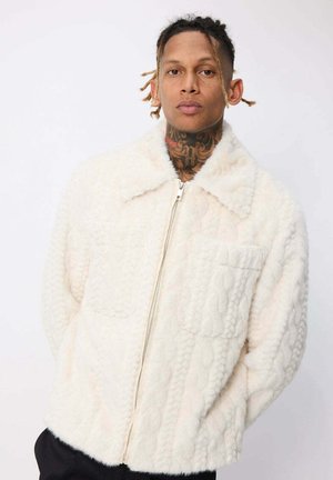 Jeune homme avec des cheveux tressés et des tatouages au cou portant une veste zippée blanche texturée avec des poches sur le devant, se tenant contre un fond uni.