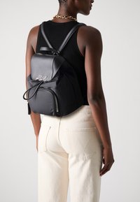 Black nylon backpack with a rounded shape, featuring a front zipper pocket, cord closure, and signature embroidery in silver.