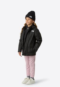 Černá voděodolná bunda s kapucí, zapínání na zip, dvě přední kapsy, nošena přes černý top. Kombinována s růžovými kalhotami a bílými teniskami.