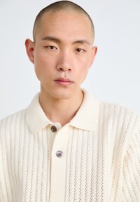 Cream-colored knitted polo shirt with vertical ribbed texture, short collar, and two brown buttons at the neckline.
