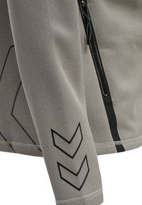 Chaqueta atlética gris que presenta una tela texturizada, mangas largas, un bolsillo con cremallera y acentos geométricos negros en la manga.