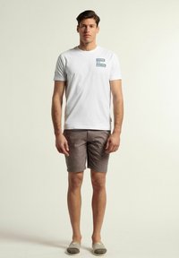 Camiseta de algodón blanca con el texto azul "GAME SET MATCH" en el pecho, combinada con pantalones cortos marrones y zapatillas grises claras de tipo slip-on.