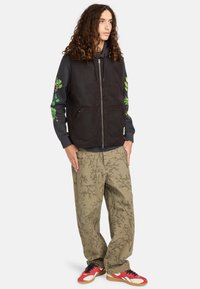 Schwarze Kapuzenweste über einem grauen Hoodie mit grünen Blättern, kombiniert mit beigen Hosen mit Astmustern und roten Sneakers.