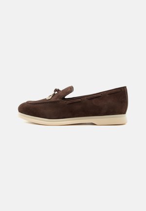 Mocassin en daim marron foncé avec semelle crème, surpiqûres latérales et pompon décoratif sur le dessus, présenté en vue de profil sur fond uni.