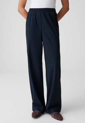 Person trägt dunkelblaue, weit geschnittene Hose mit elastischem Bund, kombiniert mit dunkelbraunen Hausschuhen und einem weißen ärmellosen Oberteil.