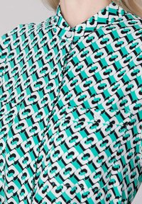 Chemise turquoise à boutons avec un motif de chaînette noir et blanc et des poches poitrine visibles.