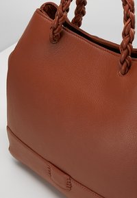 Bruine leren handtas met een gestructureerd oppervlak, voorzien van gevlochten handvatten en een gestructureerde vorm. Het heeft een decoratieve stiknaad aan de voorkant.