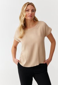 Beige short-sleeve blouse with a round neckline, soft texture, and relaxed fit. Features rolled sleeves and a slightly curved hemline.