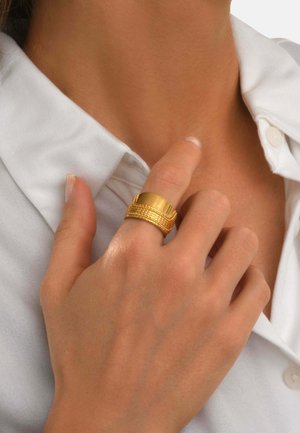 Main avec une large bague dorée texturée touchant le cou, portant une chemise blanche à col.