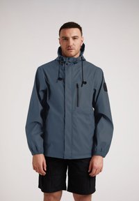 Chaqueta impermeable en azul oscuro con capucha, cordones ajustables, bolsillo en el pecho con cremallera y puños elásticos, diseñada para el uso al aire libre.